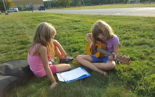 two girls sitting on the grass playing a guitar